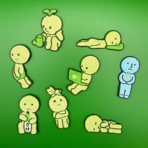 Op de afbeelding: Een verzameling geborduurde patches met grillige, cartoonachtige figuren in verschillende poses. De figuren zijn voornamelijk lichtgroen met zwarte details, en één is blauw. De patches bevinden zich op een effen groene achtergrond.