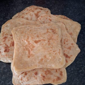 May include: A stack of golden-brown, square-shaped flatbreads. The bread has a slightly textured surface with darker brown spots, suggesting it's been cooked. The flatbreads are stacked on top of each other, with some edges visible.