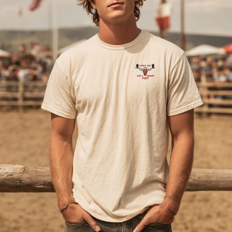 Puede incluir: Camiseta color crema con cuello redondo y mangas cortas. La camiseta presenta un peque&ntilde;o gr&aacute;fico en el pecho con las palabras "LOVELY DAY" encima de un vaso de cerveza y la frase "FOR A CREAMY PINT" debajo. La persona lleva vaqueros.
