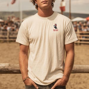 May include: Cream-coloured short-sleeved t-shirt featuring a small graphic of a person riding a barrel and the text "HOLD ON TO YOUR PINTS" in red. The shirt is made of a soft material and is suitable for casual wear.