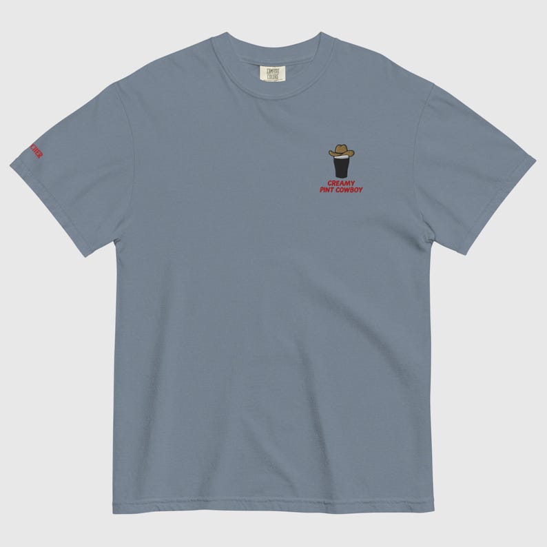 Op de afbeelding: Lichtblauw T-shirt met een geborduurd ontwerp van een cowboyhoed op een pintglas. De tekst "CREAMY PINT COWBOY" is rood. Het shirt heeft een ronde hals en korte mouwen, ideaal voor casual wear.