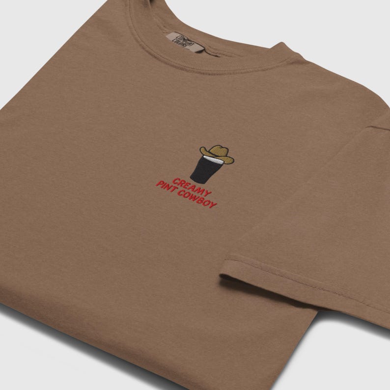 Op de afbeelding: Bruin t-shirt met een geborduurd ontwerp van een pint stout met een cowboyhoed. De tekst "CREAMY PINT COWBOY" is rood geborduurd onder het ontwerp. Het shirt heeft een ronde hals en korte mouwen.