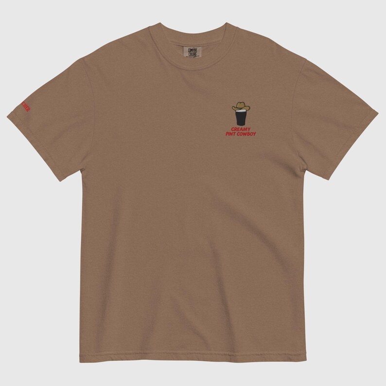Op de afbeelding: Bruin t-shirt met een kleine afbeelding van een pint bier met een cowboyhoed. De tekst "CREAMY PINT COWBOY" staat eronder in het rood. Het shirt heeft een ronde hals en korte mouwen.