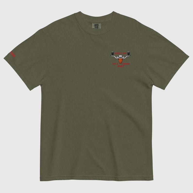 Puede incluir: Camiseta verde oliva con un peque&ntilde;o gr&aacute;fico de un toro con sombrero de vaquero y dos jarras de cerveza. El texto "Sip the Legend" y "For a Legendary Pint" est&aacute; debajo. Un peque&ntilde;o logotipo rojo est&aacute; en la manga izquierda.