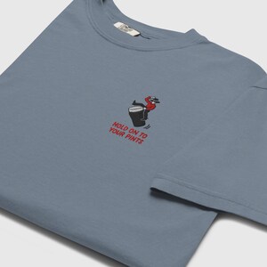 May include: A blue t-shirt featuring an embroidered design of a person on a beer mug, with the text "HOLD ON TO YOUR PINTS". The shirt has a crew neck and short sleeves.