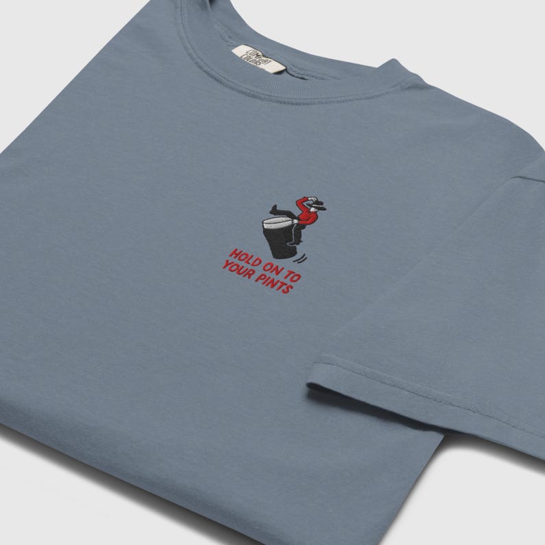 May include: A blue t-shirt featuring an embroidered design of a person on a beer mug, with the text "HOLD ON TO YOUR PINTS". The shirt has a crew neck and short sleeves.