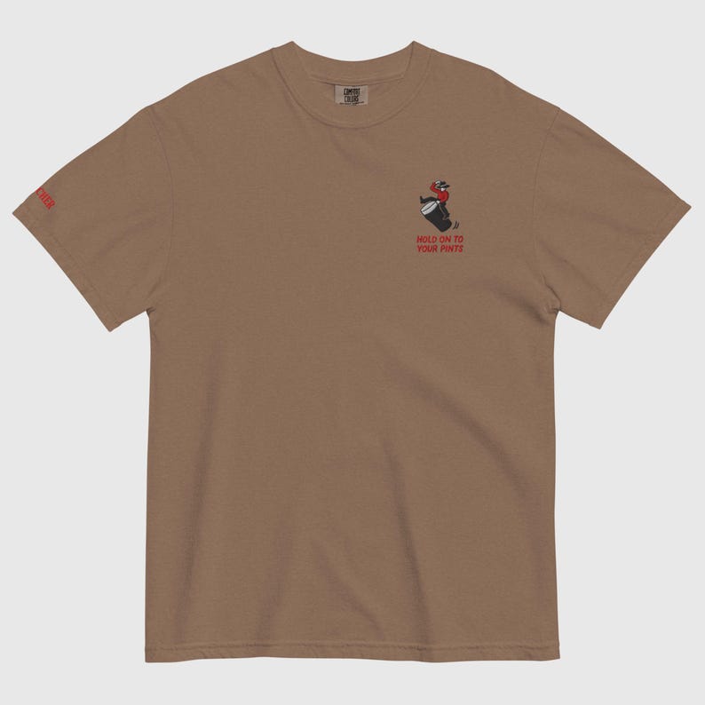 May include: A brown t-shirt featuring a cartoon figure holding a barrel, with the words "HOLD ON TO YOUR PINTS" beneath. The left sleeve displays the word "BEER".