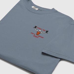 Puede incluir: Camiseta azul polvoriento con cuello redondo. La camiseta presenta un dise&ntilde;o bordado de un toro de cuernos largos con sombrero de vaquero, sosteniendo dos pintas de cerveza, y el texto "LOVELY DAY FOR A CREAMY PINT."