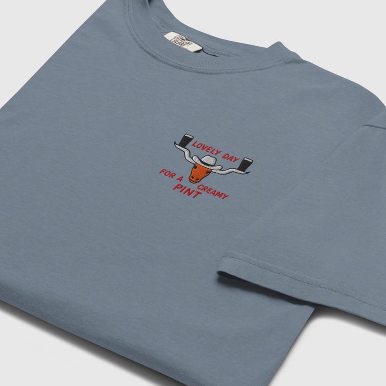 Puede incluir: Camiseta azul polvoriento con cuello redondo. La camiseta presenta un dise&ntilde;o bordado de un toro de cuernos largos con sombrero de vaquero, sosteniendo dos pintas de cerveza, y el texto "LOVELY DAY FOR A CREAMY PINT."