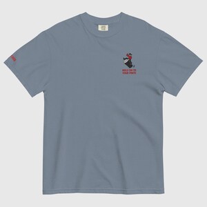 May include: A blue t-shirt with a small embroidered graphic of a person riding a barrel, with the text "HOLD ON TO YOUR PINTE" below. The word "CHER" is printed on the left sleeve in red. The shirt has short sleeves.