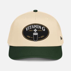 Puede incluir: Gorra de b&eacute;isbol color crema con visera y bot&oacute;n verde oscuro. La parte delantera presenta un parche ovalado bordado con las palabras "VITAMIN G" sobre un vaso de l&iacute;quido oscuro. El parche est&aacute; bordeado en dorado, con espigas de trigo a cada lado.