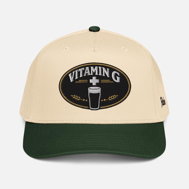 Puede incluir: Gorra de b&eacute;isbol color crema con visera y bot&oacute;n verde oscuro. La parte delantera presenta un parche ovalado bordado con las palabras "VITAMIN G" sobre un vaso de l&iacute;quido oscuro. El parche est&aacute; bordeado en dorado, con espigas de trigo a cada lado.