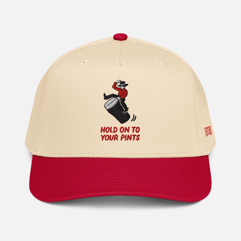May include: A cream and red baseball cap with the text "HOLD ON TO YOUR PINTS". The cap has an embroidered design of a person riding a pint glass. The cap has a red button on top.