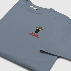 Op de afbeelding: Een stoffig blauw t-shirt met een kleine afbeelding van een pint stout met een cowboyhoed. De tekst "CREAMY PINT COWBOY" is in het rood onder de afbeelding gedrukt. Het shirt heeft een ronde hals en korte mouwen.