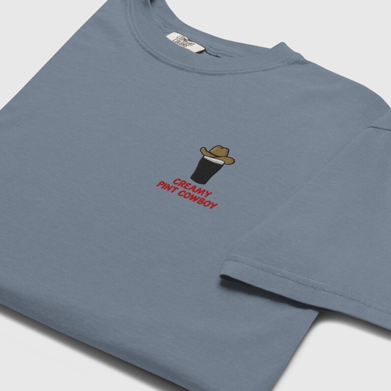 Op de afbeelding: Een stoffig blauw t-shirt met een kleine afbeelding van een pint stout met een cowboyhoed. De tekst "CREAMY PINT COWBOY" is in het rood onder de afbeelding gedrukt. Het shirt heeft een ronde hals en korte mouwen.