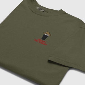 Op de afbeelding: Olijfgroen t-shirt met een geborduurd ontwerp van een pint stout met een cowboyhoed. De tekst "CREAMY PINT COWBOY" is in het rood geborduurd. Het shirt heeft een ronde hals en korte mouwen.