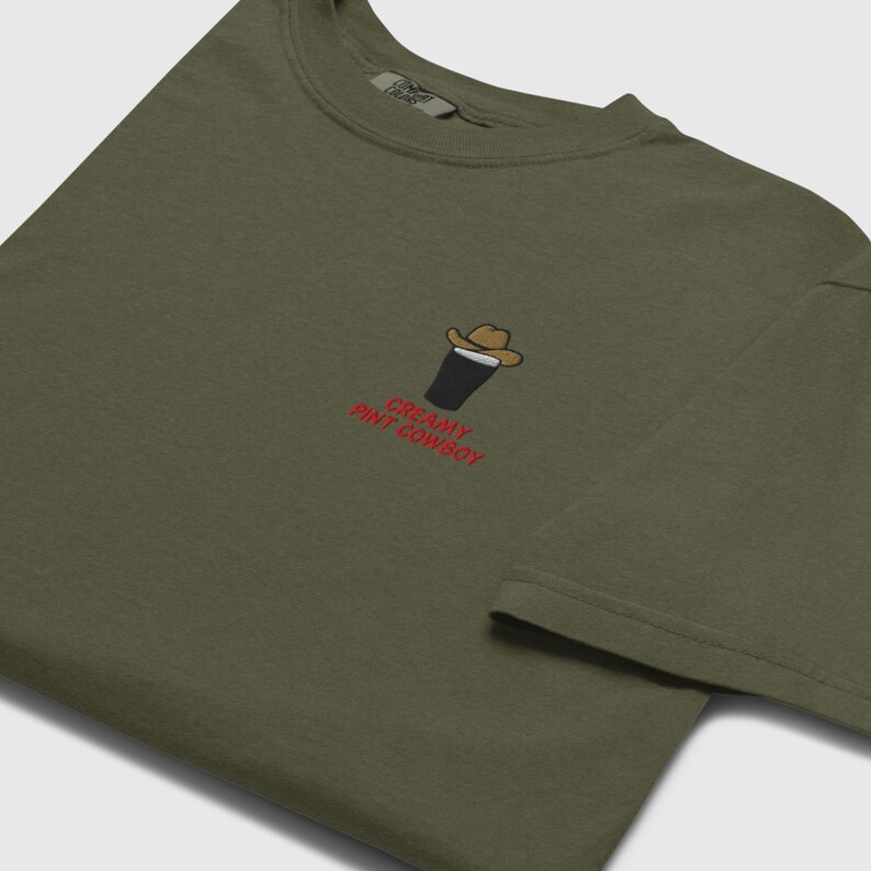 Op de afbeelding: Olijfgroen t-shirt met een geborduurd ontwerp van een pint stout met een cowboyhoed. De tekst "CREAMY PINT COWBOY" is in het rood geborduurd. Het shirt heeft een ronde hals en korte mouwen.
