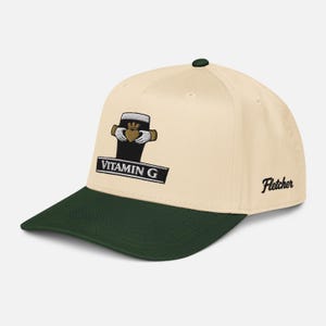 May include: Cream-colored baseball cap with a dark green brim and top button. The cap features an embroidered design of a pint of beer with a heart and hands, and the text "Vitamin G". The word "Fletcher" is embroidered on the side.