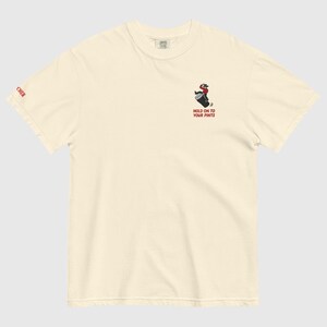 May include: Off-white t-shirt featuring a cartoon cowboy riding a barrel, with the text "HOLD ON TO YOUR PINTS" in red. The word "CHER" is printed in red on the left sleeve. The shirt is made of a soft, comfortable material.