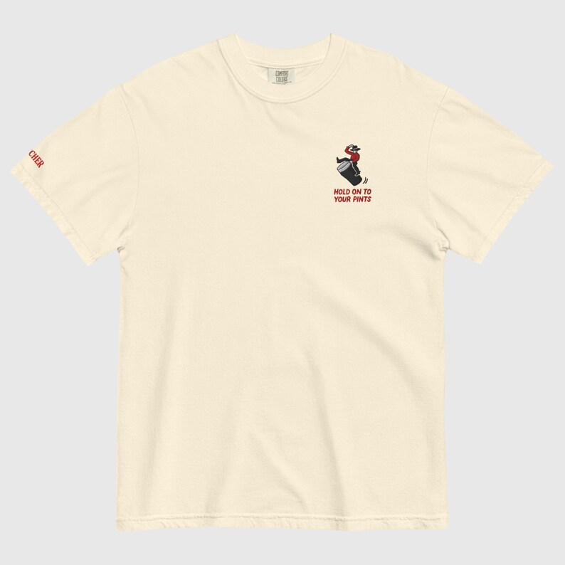 May include: Off-white t-shirt featuring a cartoon cowboy riding a barrel, with the text "HOLD ON TO YOUR PINTS" in red. The word "CHER" is printed in red on the left sleeve. The shirt is made of a soft, comfortable material.