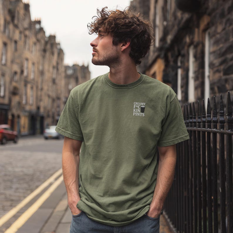 K&ouml;nnte beinhalten: Olivgr&uuml;nes T-Shirt mit dem Text "CREAMY F*CKIN PINTS" und einer kleinen Grafik eines Bierkrugs. Das Shirt hat kurze &Auml;rmel und einen Rundhalsausschnitt. Die Person steht auf einer Stra&szlig;e in der Stadt.
