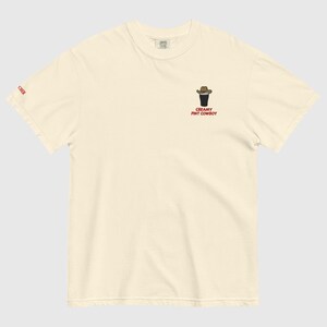 Op de afbeelding: Cr&egrave;mekleurig T-shirt met een kleine afbeelding van een pintglas met een cowboyhoed en de tekst "CREAMY PINT COWBOY" in het rood. Het woord "CHER" staat in het rood op de linkermouw. Het shirt is gemaakt van zacht materiaal.