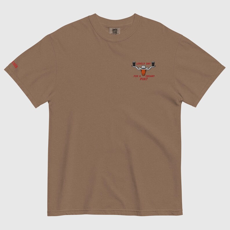 Puede incluir: Camiseta marr&oacute;n con un gr&aacute;fico de dos pintas de cerveza y el texto "Lovely Day for a Creamy Pint". La palabra "Cheer" est&aacute; impresa en la manga izquierda. La camiseta est&aacute; hecha de un material suave.