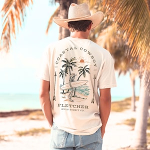May include: Cream-colored t-shirt with a graphic of a skeleton surfer carrying a surfboard on a beach with palm trees. The text "Coastal Cowboy" arches across the top, with "Fletcher Gulf & Grit Co." below.