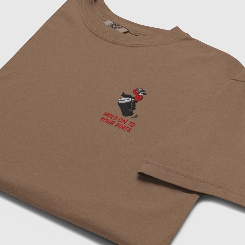 May include: A brown t-shirt featuring an embroidered graphic of a person on a beer mug. The text "HOLD ON TO YOUR PINTS" is in red. The shirt has a relaxed fit and a crew neck.