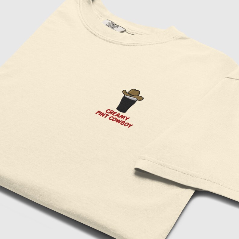 Op de afbeelding: Cr&egrave;mekleurig T-shirt met een kleine geborduurde afbeelding van een pint stout met een cowboyhoed. De tekst "Creamy Pint Cowboy" is eronder in het rood geborduurd. Het shirt heeft een ronde hals en korte mouwen.