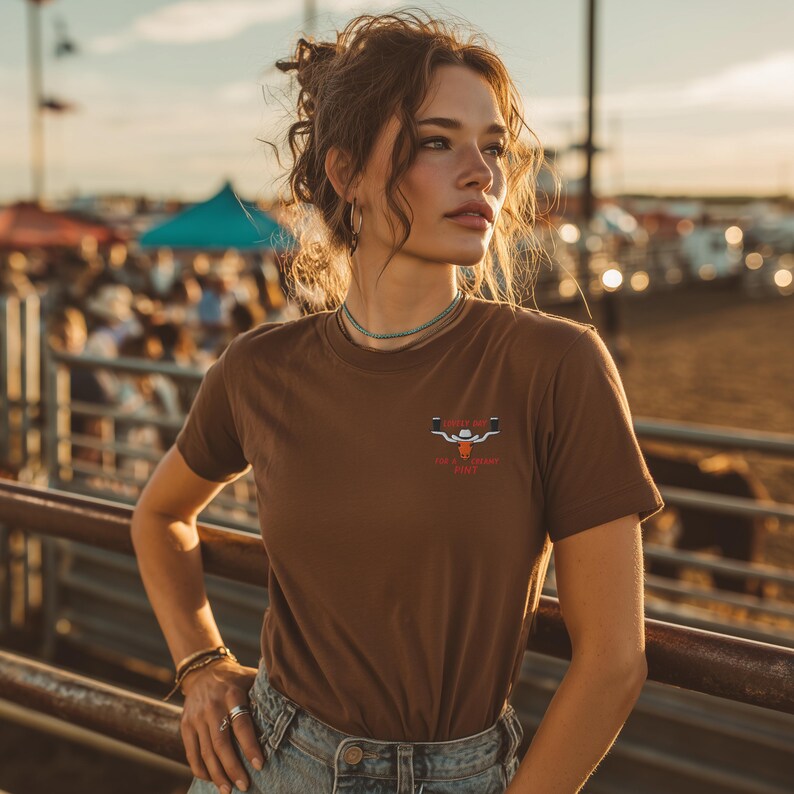 Puede incluir: Una camiseta marr&oacute;n con un gr&aacute;fico de una cabeza de toro y el texto "Cowboy Up" en rojo. La camiseta de manga corta est&aacute; hecha de un material suave. La camiseta la lleva una persona.