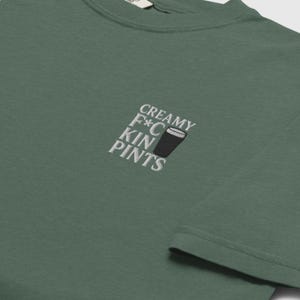 K&ouml;nnte beinhalten: Gr&uuml;nes T-Shirt mit der gestickten Aufschrift "CREAMY F*C KIN PINTS" und einer Abbildung eines Bierkrugs. Das Shirt hat einen Rundhalsausschnitt und ist aus weichem Stoff gefertigt.