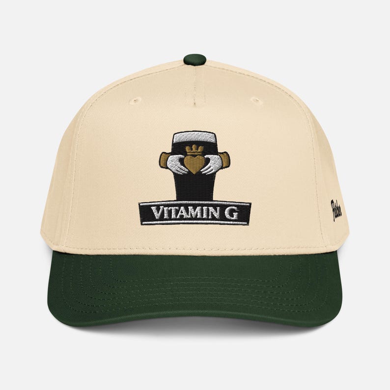 May include: A baseball cap with a cream-colored crown and a dark green brim. The cap features an embroidered design of a black pint glass with a gold crown and hands, and the text "VITAMIN G" in a black rectangle.