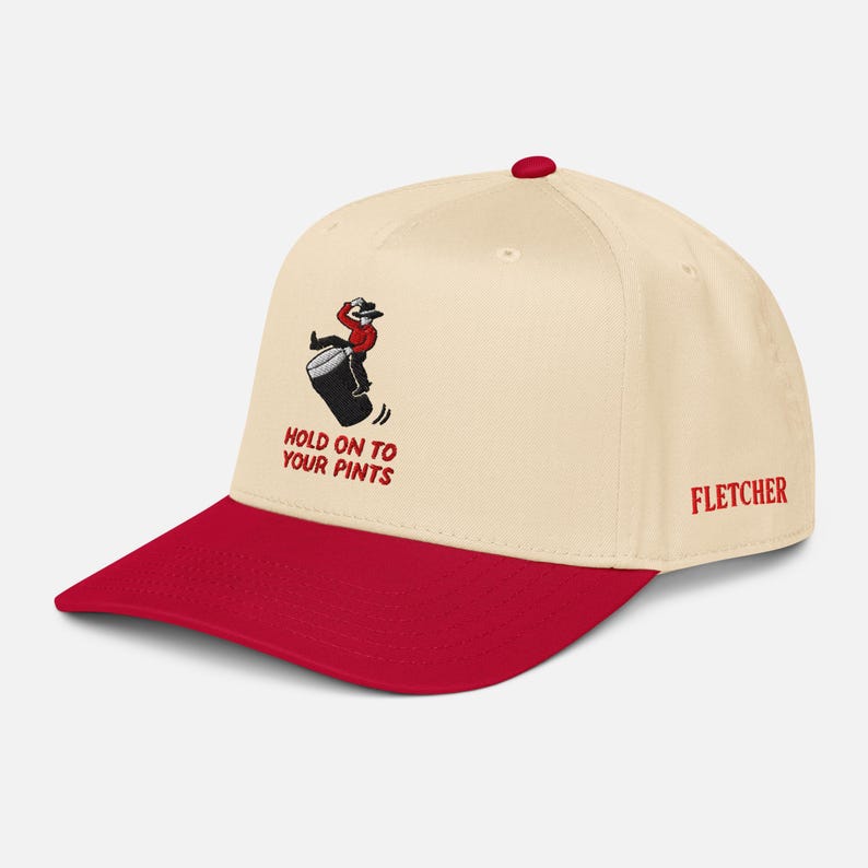 May include: Baseball cap in cream with a red peak and button. It has an embroidered graphic of a person falling off a barrel, with the text "HOLD ON TO YOUR PINTS." The name "FLETCHER" is embroidered on the side.