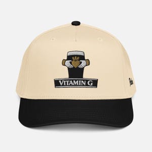 May include: Cream-colored baseball cap with a black brim. The cap features an embroidered design of a black pint glass with a gold crown and hands holding a heart. The text "VITAMIN G" is below the design.