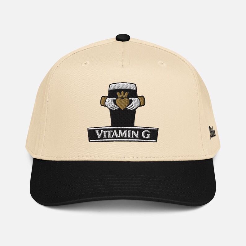May include: Cream-colored baseball cap with a black brim. The cap features an embroidered design of a black pint glass with a gold crown and hands holding a heart. The text "VITAMIN G" is below the design.
