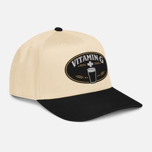 Puede incluir: Gorra de b&eacute;isbol color crema con visera negra. La gorra presenta un parche ovalado bordado con las palabras "VITAMIN G" y un gr&aacute;fico de bebida. El parche est&aacute; contorneado en dorado.