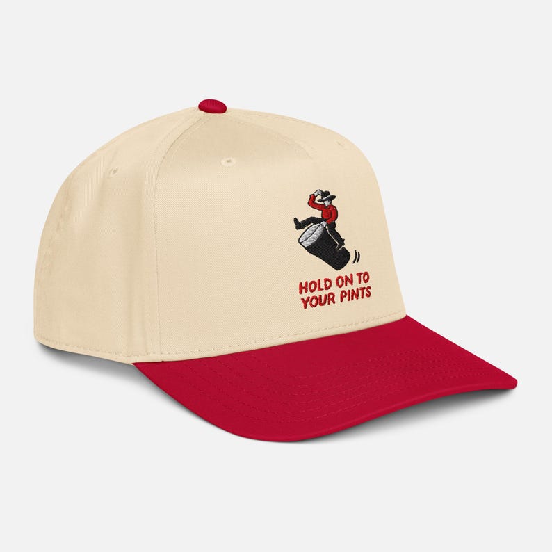 May include: A cream and red baseball cap with the text "HOLD ON TO YOUR PINTS". The cap has an embroidered design of a person riding a barrel. The brim and button are red.