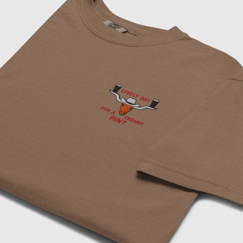 Puede incluir: Camiseta marr&oacute;n con un gr&aacute;fico de un longhorn con sombrero de vaquero y dos pintas de cerveza. El texto dice "LOVELY DAY FOR A CREAMY PINT" en rojo y blanco.