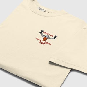 Puede incluir: Camiseta de color crema con un dise&ntilde;o bordado. El dise&ntilde;o incluye una cabeza de toro con sombrero de vaquero, con el texto "LOVELY DAY FOR A CREAMY PINT" en rojo. La camiseta tiene cuello redondo y mangas cortas.