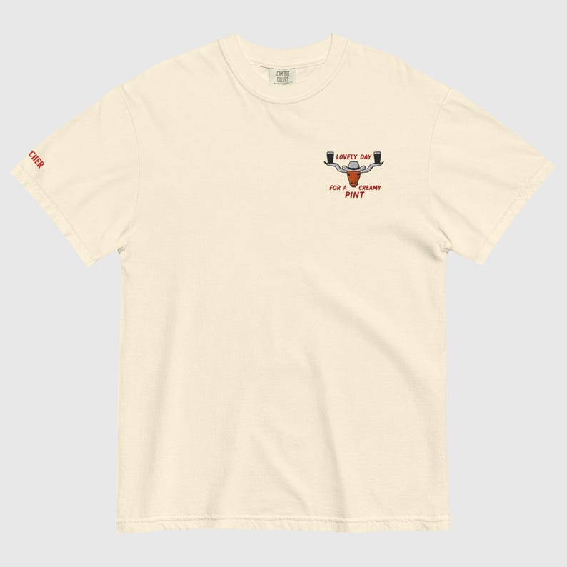 Puede incluir: Camiseta de color crema con un peque&ntilde;o gr&aacute;fico de una cabeza de toro con dos jarras de cerveza y el texto "LOVELY DAY FOR A CREAMY PINT". La palabra "CHER" est&aacute; impresa en la manga izquierda.