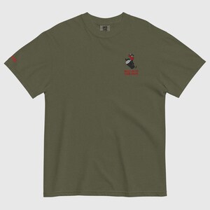 May include: Olive green t-shirt featuring a small graphic of a person playing a drum and the text "HOLD ON TO YOUR FIRE" in red. The same text is also printed on the left sleeve in red.