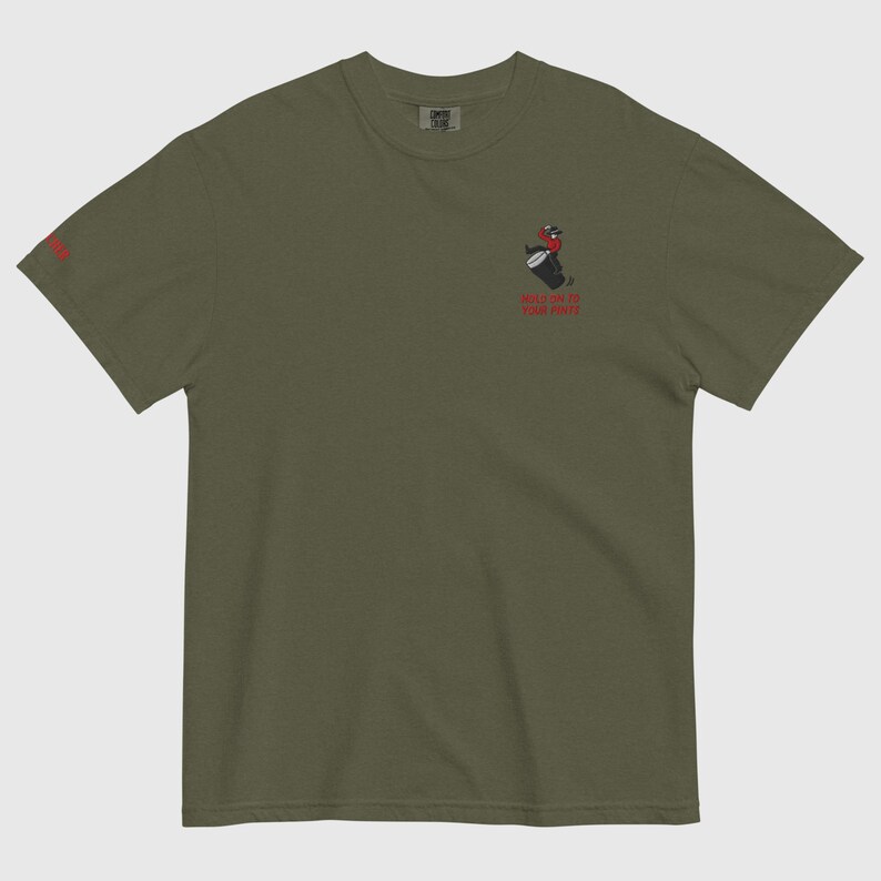 May include: Olive green t-shirt featuring a small graphic of a person playing a drum and the text "HOLD ON TO YOUR FIRE" in red. The same text is also printed on the left sleeve in red.
