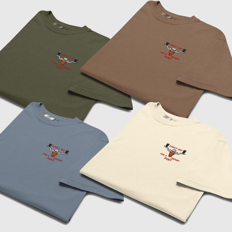 Puede incluir: Cuatro camisetas en verde oliva, marr&oacute;n, azul polvoriento y crema. Cada camiseta presenta un peque&ntilde;o dise&ntilde;o bordado de una cabeza de toro con dos vasos de cerveza y el texto "LOVELY DAY FOR A CREAMY PINT" en rojo.