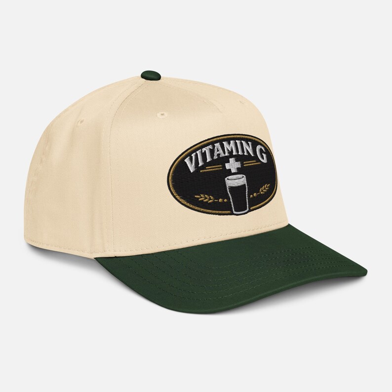 Puede incluir: Gorra de b&eacute;isbol color crema con visera y bot&oacute;n verde oscuro. La gorra presenta un parche ovalado bordado con las palabras "VITAMIN G" sobre un gr&aacute;fico de un vaso y una cruz.