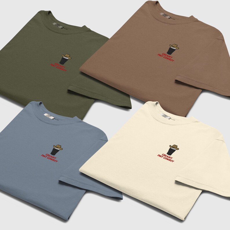 Op de afbeelding: Vier t-shirts in olijfgroen, bruin, blauw en cr&egrave;me. Elk shirt heeft een klein geborduurd ontwerp van een pintglas met een cowboyhoed, met de tekst "CREAMY PINT COWBOY" in het rood eronder.