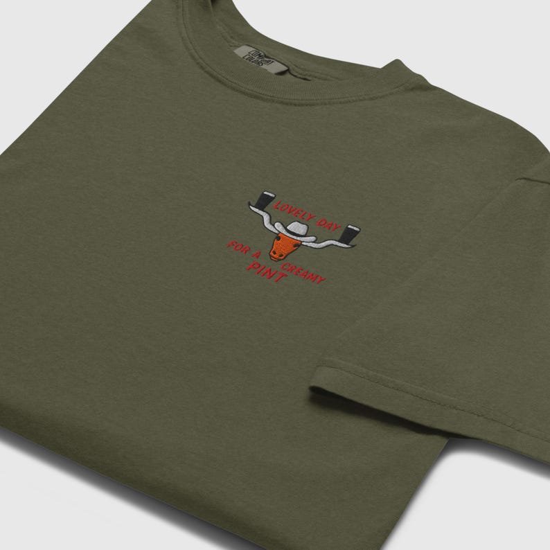 Puede incluir: Camiseta verde oliva con un dise&ntilde;o bordado. El dise&ntilde;o presenta un toro de cuernos largos con un sombrero de vaquero, sosteniendo dos pintas de cerveza y el texto "LOVELY DAY FOR A CREAMY PINT" en rojo.