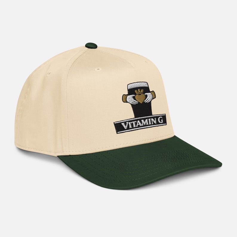 May include: Cream-colored baseball cap with a dark green brim and top button. The cap features an embroidered design of a pint of stout with a gold heart and the text "VITAMIN G" on a black rectangle.