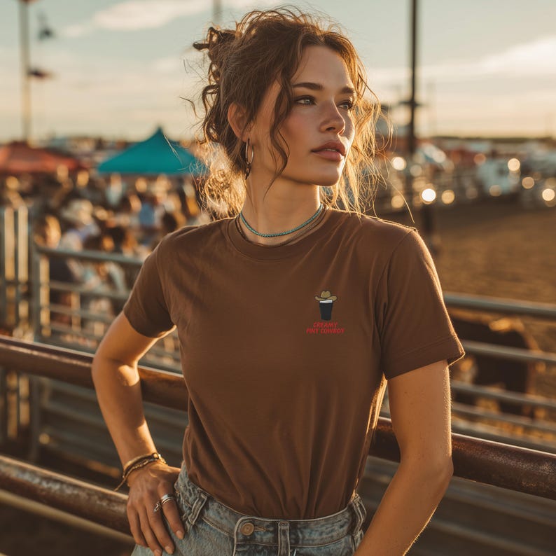 Op de afbeelding: Bruin t-shirt met een kleine afbeelding van een cowboyhoed en een drankje, met de rode tekst "TOUGH BUT TENDER". Het shirt wordt gedragen met een blauwe spijkerbroek. De achtergrond toont een rodeo of buitenevenement.