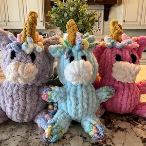 Puede incluir: Tres juguetes de peluche de unicornio de crochet. El unicornio de la izquierda es morado, el del medio es azul y el de la derecha es rosa. Cada unicornio tiene un cuerno dorado y una cara blanca.